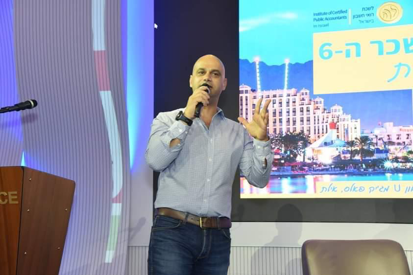 יוני ירימי מרצה מול קהל, מדבר למיקרופון על במה